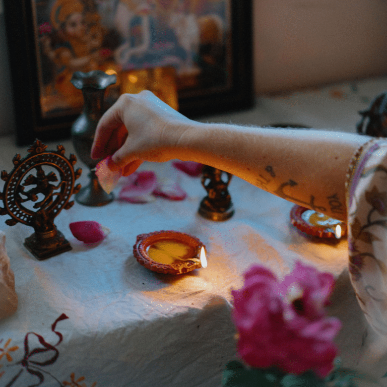 A hand reaches over to a white table cloth. The hand is holding a rose petal, and there is a pink rose and candles on the table.