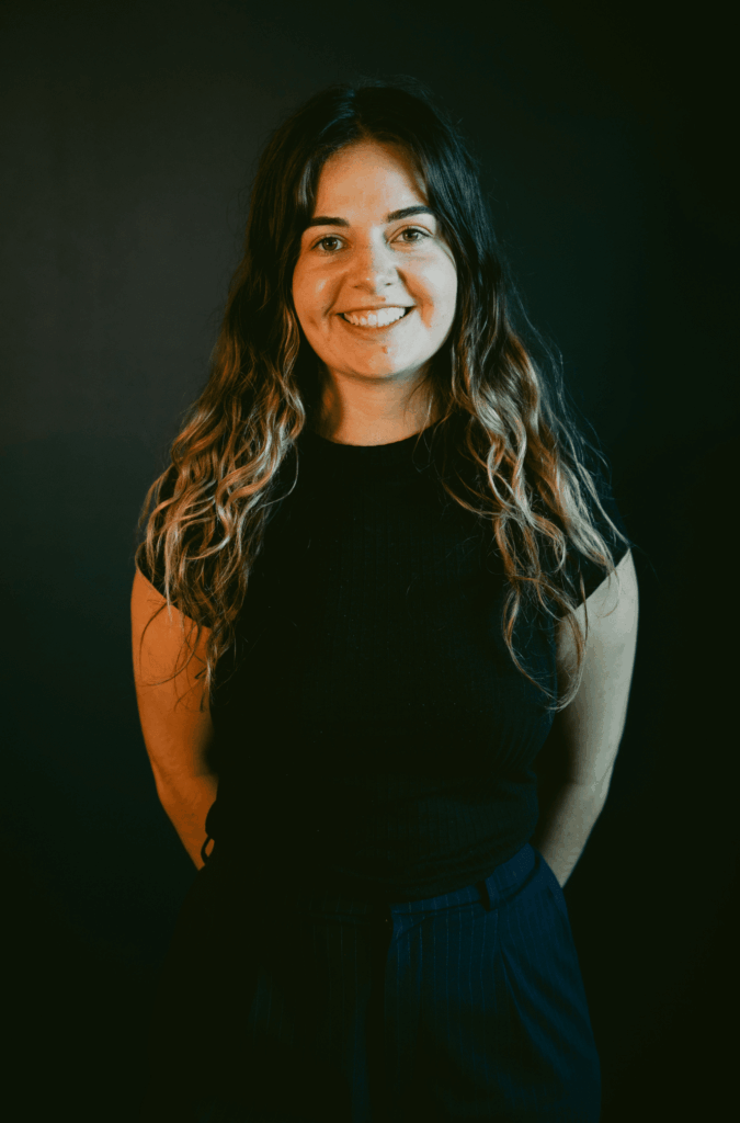 a woman with brown hair stands in front of a black wall smiling
