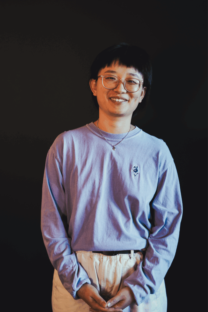 a person in a long sleeved purple shirt stands in front of a dark wall