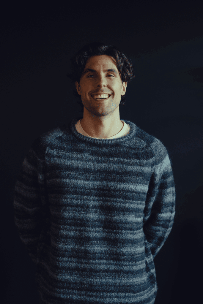 A man with brown hair in a blue striped jumper stands smiling