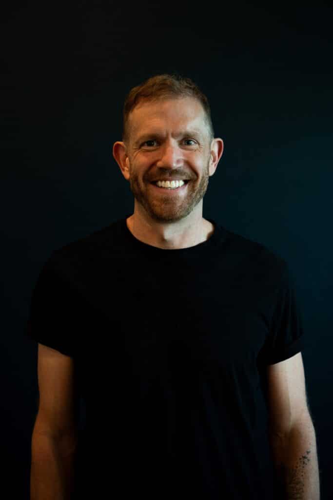 a man in a black t shirt smiles against a dark backdrop